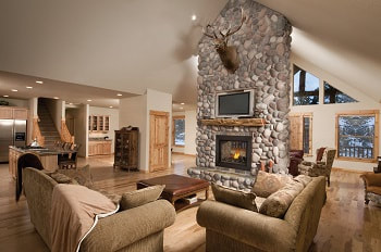 Double sided gas fireplace positioned in the middle of a living room, a loveseat and sofa out front of one fireplace side, the fireplace enclosure is finished with natural river rock stone, there is a light natural wood mantel above the fireplace opening, a tv and a stuffed deer.